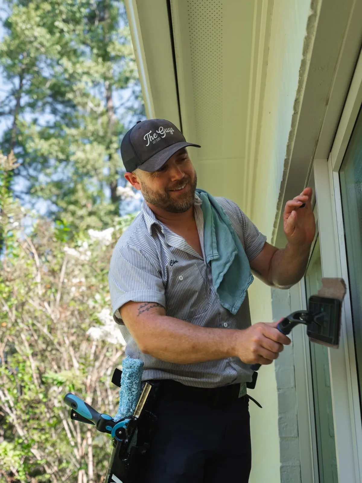 A smiling Guys window cleaner in a branded cap washing an exterior window with a soapy applicator