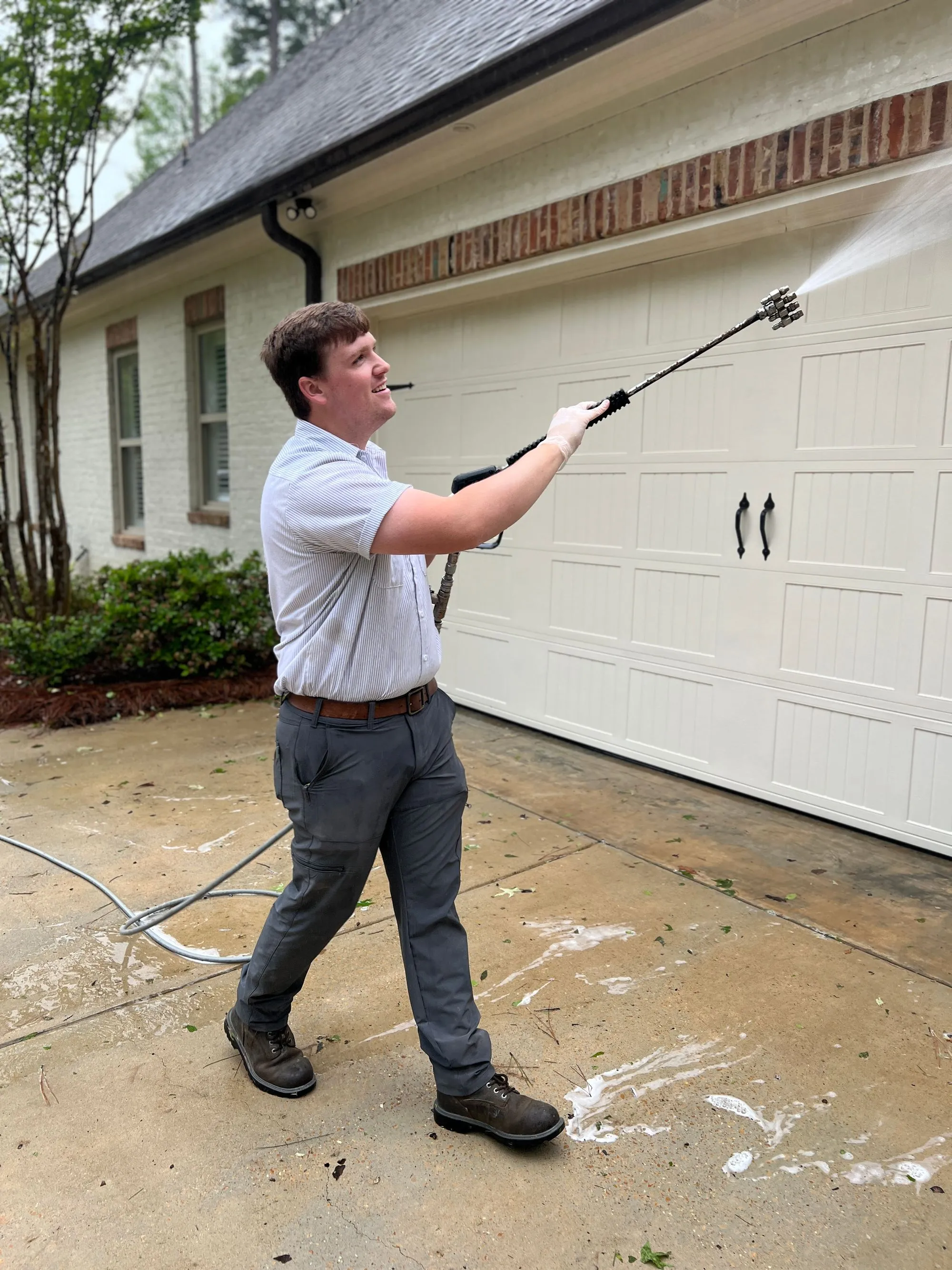 A Guys technician using a long telescoping wand to soft-wash the trim above a white brick home's garage
